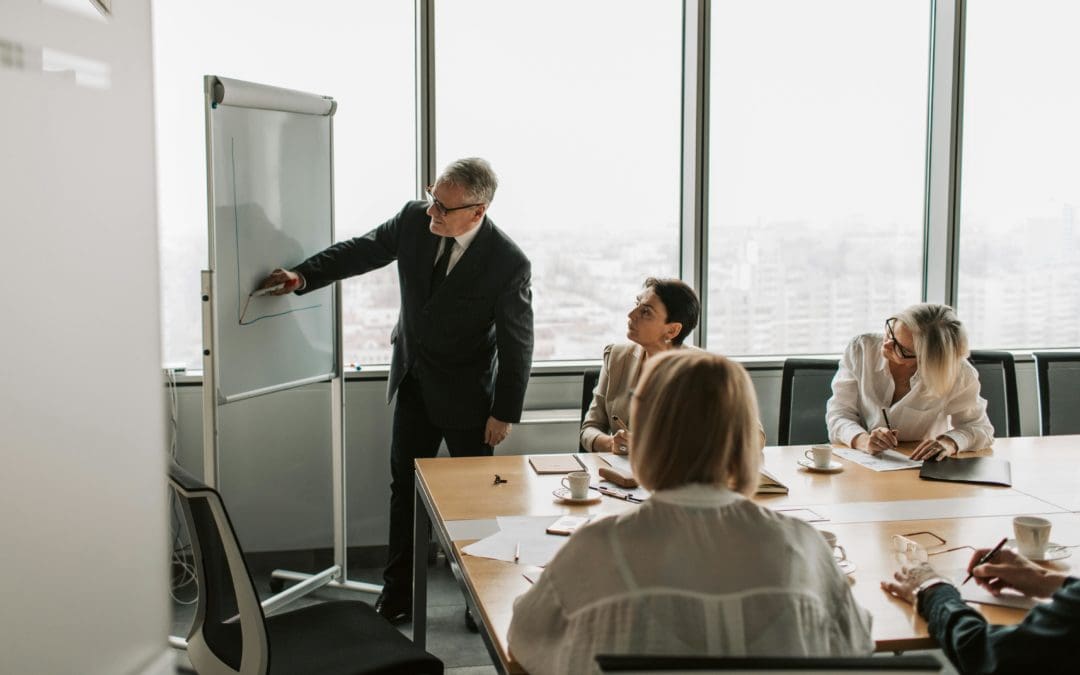 A business meeting in a modern office with a senior executive presenting a graph on a whiteboard while colleagues take notes and listen attentively.